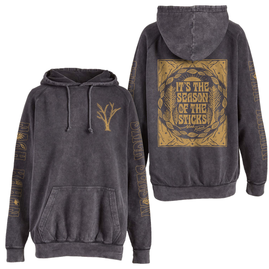 the official Noah Kahan Stick Season Hoodie in vintage black mineral wash. "It's the season of the sticks" on the back, tree branches on front, and full length prints on each sleeve, all in gold metallic ink. 